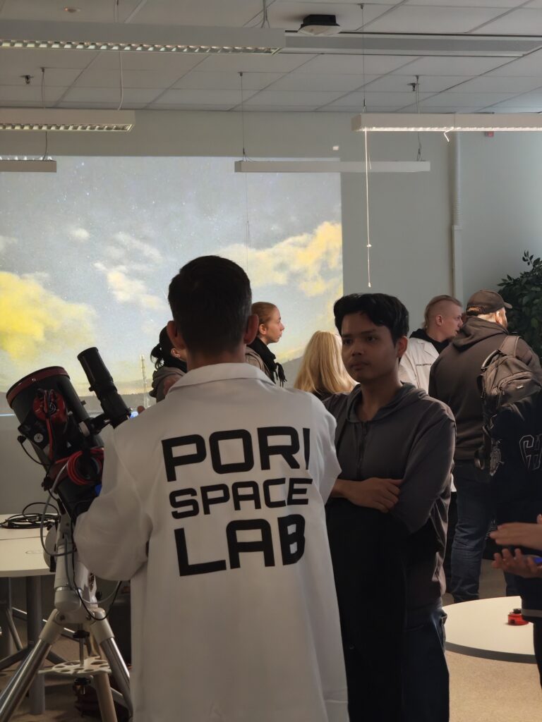 Audience at Researcher's night, a person in a lab coat with their back to the camera is standing next to a telescope, and in the background there are more people and a sky with some clouds is projected to the wall behind them.