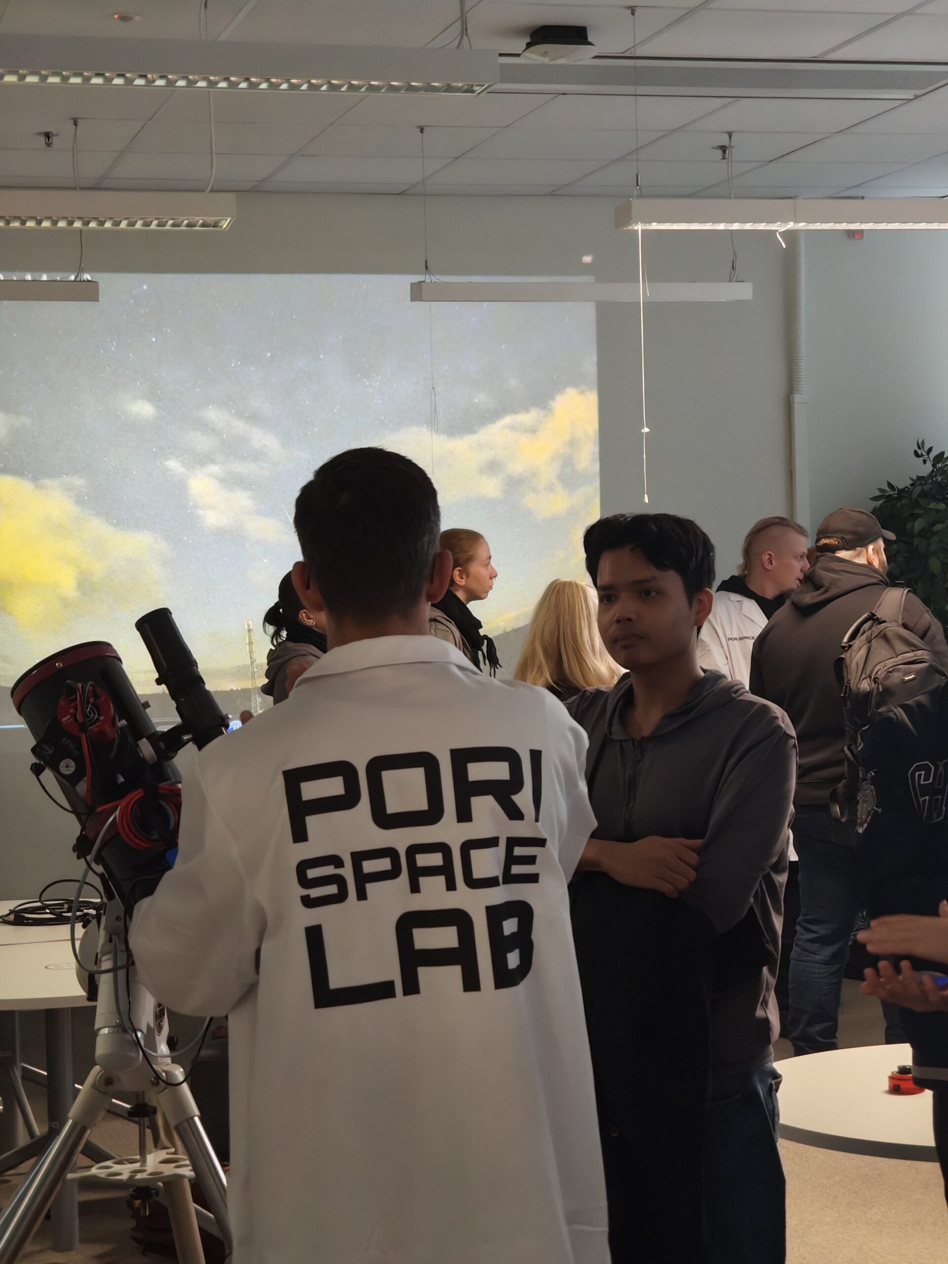 Audience at Researcher's night, a person in a lab coat with their back to the camera is standing next to a telescope, and in the background there are more people and a sky with some clouds is projected to the wall behind them.